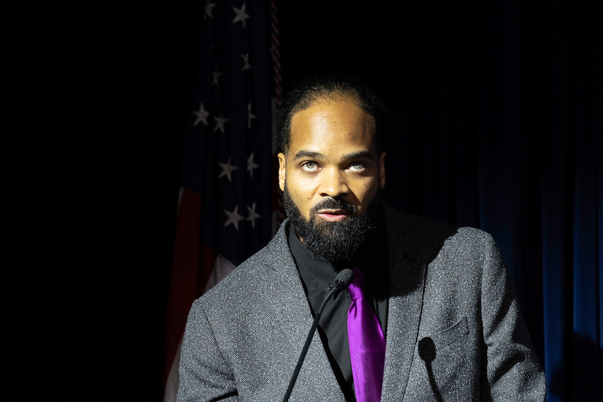 eli williamson speaking at a microphone with an American flag behind him